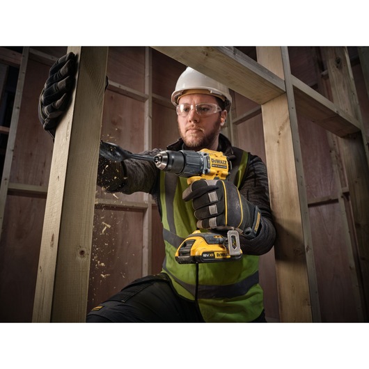 A person wearing gloves, a high-vis vest, and a hard hat is using a yellow DEWALT cordless drill (DCD799D2T) to drill into a wooden beam inside a construction site. Wood shavings are visible as the drill penetrates the wood.