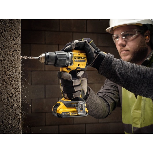 A person wearing gloves uses a DEWALT DCD799D2T 18V XR cordless drill to drill into a concrete wall. The drill is yellow and black and features the DEWALT branding.