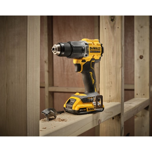 A DEWALT cordless power drill model DCD799D2T is placed on a wooden beam at a construction site. Wood shavings and a drill bit are also visible on the beam.