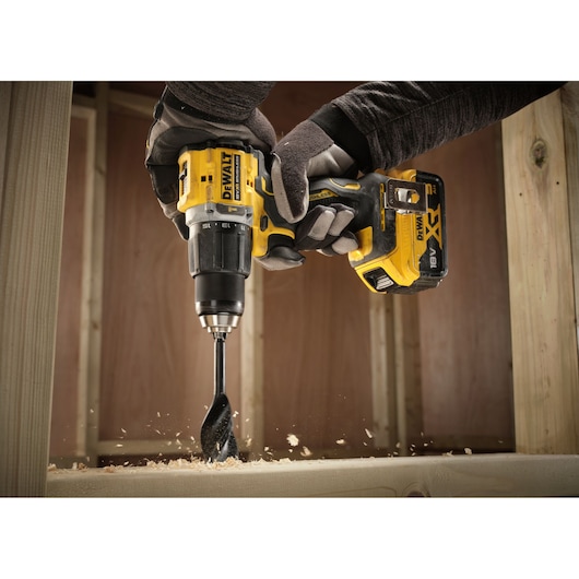A pair of gloved hands using a DEWALT cordless drill to bore a hole in a wooden beam, with wood shavings flying as the drill is in action.