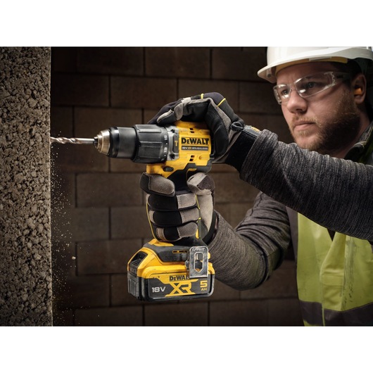 A person wearing work gloves and a safety vest is using a DEWALT 18V XR cordless drill to drill into a concrete wall. The drill is yellow and black, and dust is visible as the bit penetrates the surface.