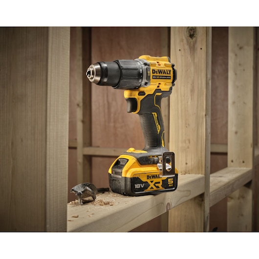 A DEWALT 18V XR brushless cordless drill is placed on a wooden beam inside an unfinished construction area. The drill has a yellow and black design and a 5Ah battery attached. Wood shavings and a drill bit are visible nearby.