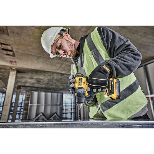 A person wearing a safety helmet, gloves, and a high-visibility vest is using a DEWALT DCD800E1T cordless drill to drill