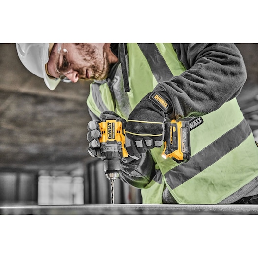 A construction worker wearing safety gear uses a yellow DEWALT cordless drill to drill into metal.