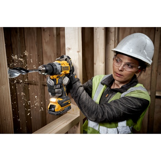 DEWALT DCD800E1T drill in use, drilling through wood with shavings flying, worker wearing gloves and helmet.