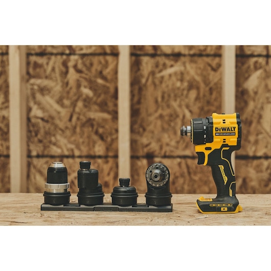 A DEWALT cordless drill driver with four interchangeable attachments displayed on a wooden surface, set against an unfinished plywood wall background.