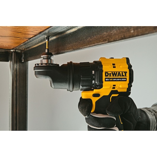 A DEWALT 18V XR Brushless drill being used with a right angle attachment to drive a screw into a metal frame under a wooden surface. The drill is held by a gloved hand.