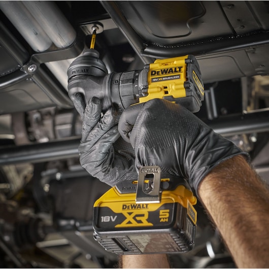 Close-up of a person wearing black gloves using a DEWALT 18V XR cordless drill with a right angle attachment to drive a screw into metal framework under a vehicle.