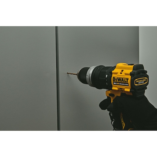 A DEWALT 18V XR brushless cordless drill being used to drill into a gray wall. The drill is held by a gloved hand and is actively engaged in the drilling process.