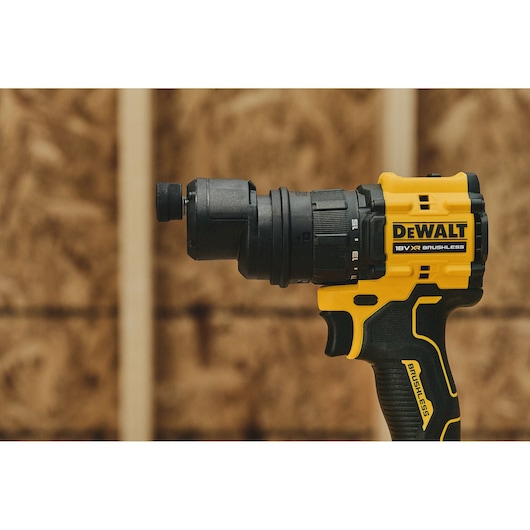 Close-up of a DEWALT 18V XR brushless cordless drill, showing the yellow and black body and the chuck. The background is a wooden wall with a blurred effect.
