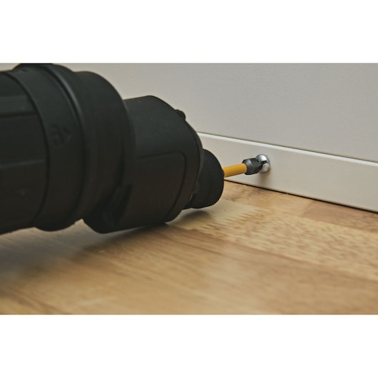 A close-up view of a power drill driving a screw into a white piece of furniture on a wooden surface.