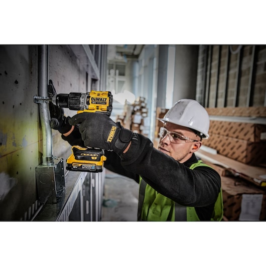 A person wearing a hard hat, safety vest, and gloves uses a DEWALT DCD805E1T power drill to work on a metal conduit attached to a wall in an industrial or construction setting.