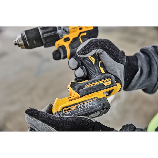 Close-up of a person wearing gloves holding a DEWALT cordless drill with a POWERSTACK 18V battery attached.