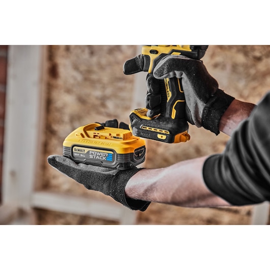 Gloved hands hold a DEWALT cordless drill with a Powerstack battery inside a workshop.
