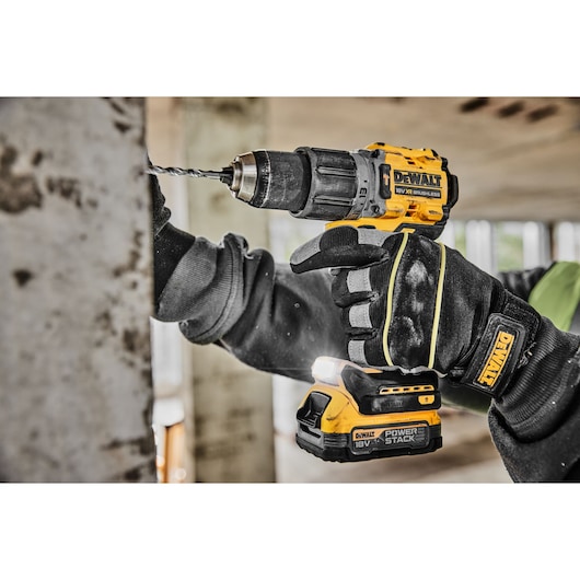 A person wearing black work gloves is using a yellow and black DEWALT cordless drill to drill into a concrete surface indoors.