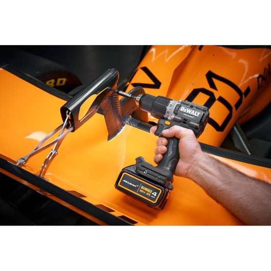 A close-up of a person using a DEWALT cordless drill on a carbon fiber automotive part with orange bodywork in the background.