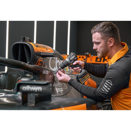 A person using a DEWALT DCD85MM1T cordless drill on the side of an orange and black race car in a workshop. The person's face is blurred for privacy, and the car has visible sponsor logos.
