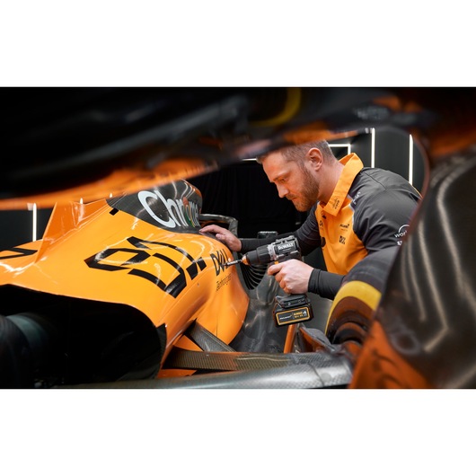 A person is using a DEWALT cordless drill to work on the body of a bright orange race car, with the car's branding visible. The individual's face is blurred for privacy.