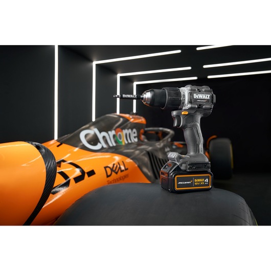 A DEWALT cordless drill is positioned on the tire of an orange Formula 1 car with McLaren and sponsor logos in a modern, illuminated garage.