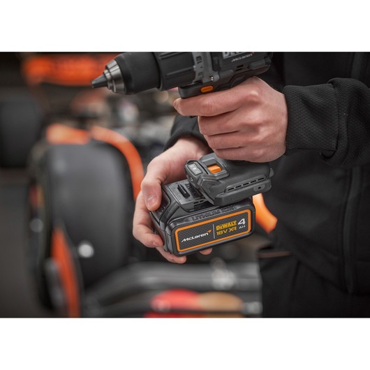 Close-up of a person holding a DEWALT 18V XR cordless power drill with a 4.0Ah battery, McLaren edition. An orange and black vehicle is blurred in the background.