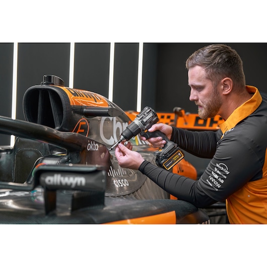 A person uses a DEWALT cordless drill on a part of a black and orange race car. The person’s face is blurred for privacy and they wear a branded team uniform.