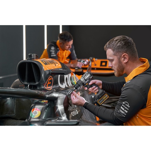 Two team members wearing orange and black uniforms are working on a race car, with one person using a DEWALT power tool on the car's exterior. The faces of both individuals are blurred for privacy.