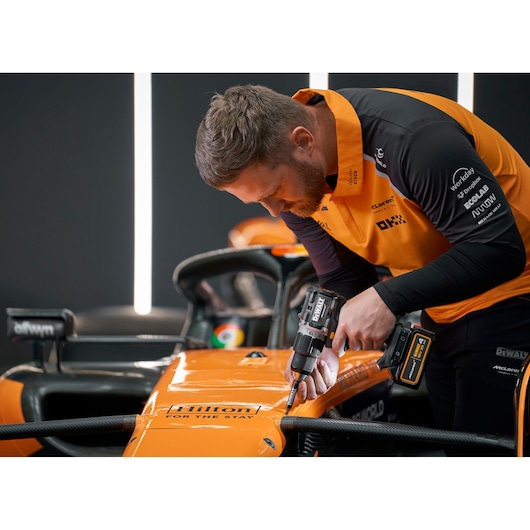 A person with their face obscured is using a DEWALT power drill to work on an orange race car in a professional garage or workshop setting. The person is wearing an orange and black uniform with various logos visible.