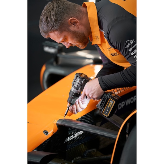 Person using a DEWALT DCD85MP2T drill on an orange McLaren car body, close-up of hands and tool.