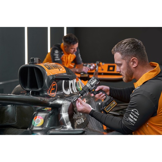 Person uses a DEWALT power drill on an orange race car. Faces are blurred and team shirts are visible.