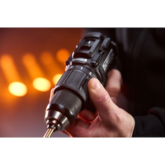 Hand holding a DEWALT cordless drill with adjustable torque, close-up with blurred orange lights in background.