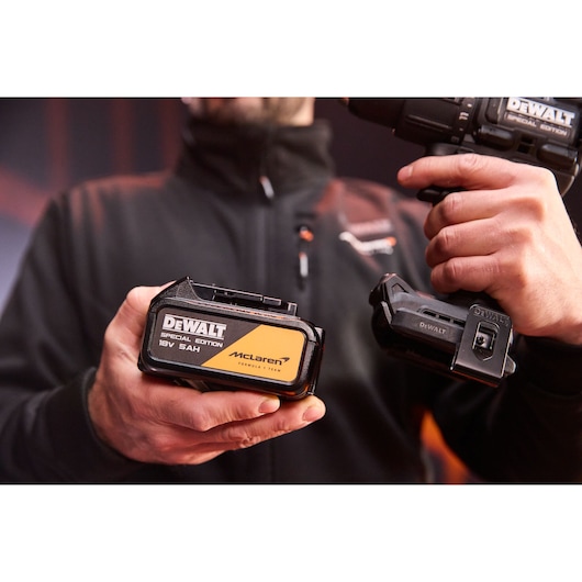 Man holds DEWALT Special Edition 18V 5Ah McLaren battery and drill against a blurred background.