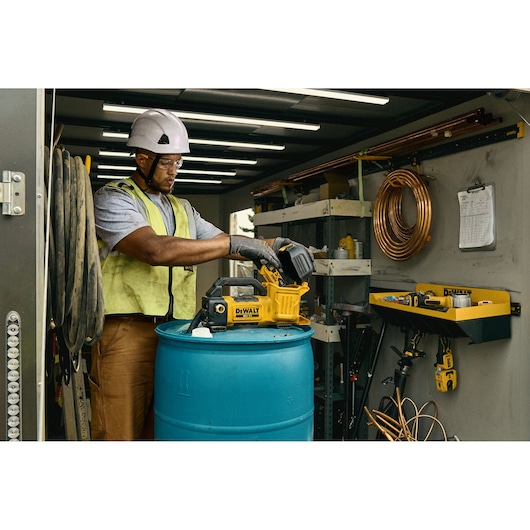 A worker wearing a hard hat and safety vest uses a DEWALT DCE050N photography-related device placed on a large blue barrel inside a well-organized workshop with various tools and coils visible on shelves and walls.
