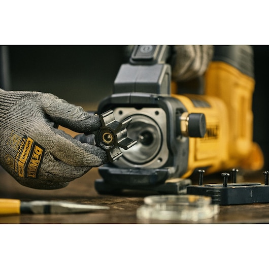 A close-up view of a gloved hand holding a tool part in front of a DEWALT photography device, with other components and tools visible on the work surface.