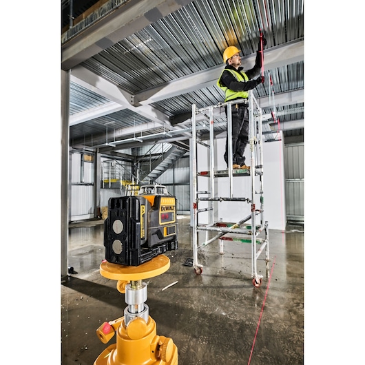 DEWALT laser level device placed on a tripod in the foreground of an industrial construction site. In the background, a worker wearing a safety helmet and vest is standing on a scaffold, using the laser line along a metal ceiling beam.
