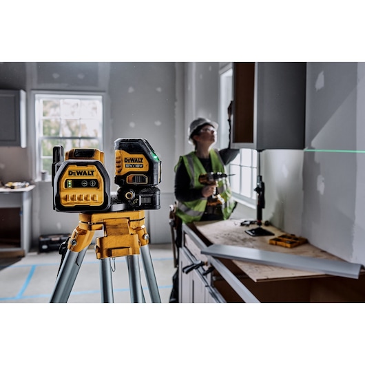 A DEWALT green line laser level (model DCE088D1G18) mounted on a tripod is positioned in a partially constructed room. A person wearing a safety helmet and vest is working in the background, using a power tool near a window. Green laser lines are visible on the wall.