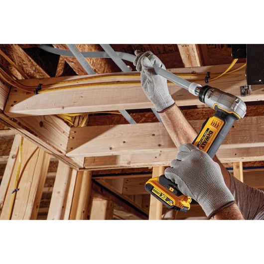 A person wearing gray work gloves uses a DEWALT cordless pipe crimping tool to secure a gray plastic pipe to wooden framing during construction.