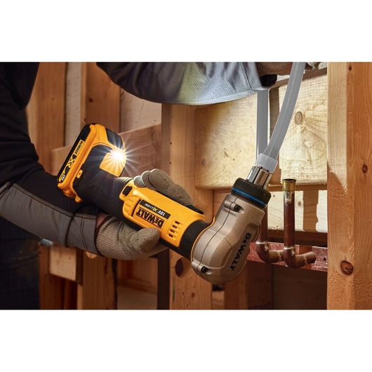 A person wearing work gloves uses a DEWALT DCE400D2 18V cordless pipe crimping tool to connect a pipe inside a wooden framed construction area.