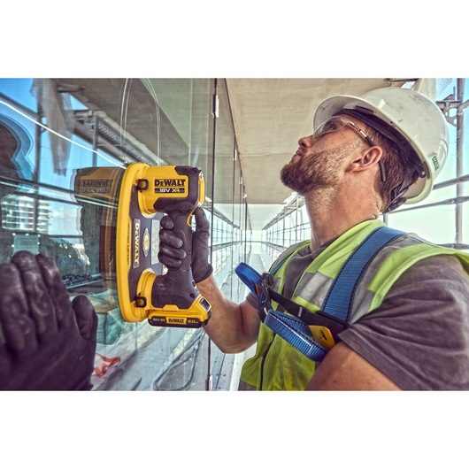 Worker uses DEWALT DCE590 tool on glass. Wearing safety gear and helmet at a construction site.