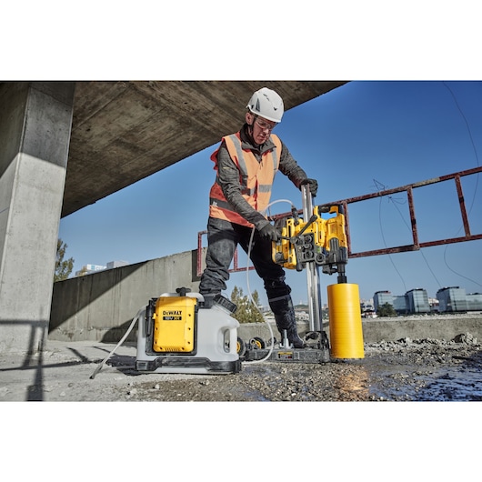 A person wearing protective gear and an orange safety vest is operating a DEWALT concrete drilling machine outdoors at a construction site. The DEWALT equipment, including a yellow drill and a white water tank with the DEWALT logo, is positioned on a concrete surface under an unfinished structure.