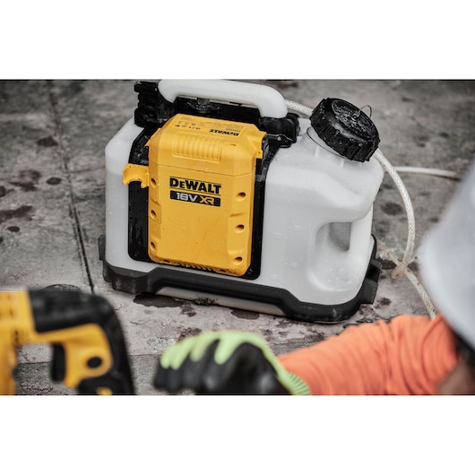 A DEWALT 18V XR cordless sprayer placed on a concrete surface. The sprayer features a white tank with a black cap and yellow battery compartment. Part of a person wearing gloves and protective gear is visible in the foreground.