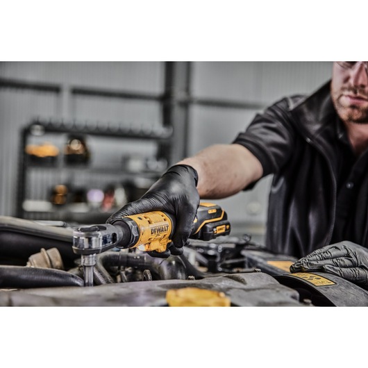A person wearing black gloves uses a yellow DEWALT cordless ratchet tool to work on a car engine in a workshop setting.
