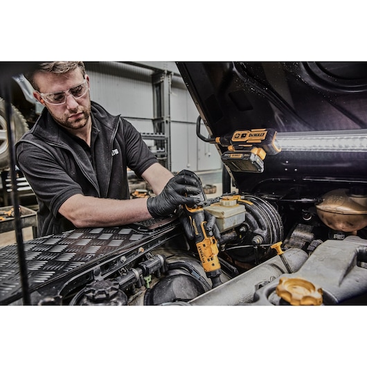 A person wearing black gloves uses a DEWALT DCF503L1G cordless ratchet tool to work on the engine of a vehicle. The vehicle hood is open, and a DEWALT work light is attached to the hood to illuminate the workspace. The person's face is blurred for privacy.