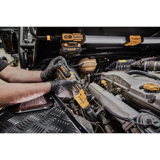 A person wearing black gloves uses a DEWALT cordless ratchet tool to work on a car engine. Additional DEWALT power tools and accessories are visible attached to the car's hood.