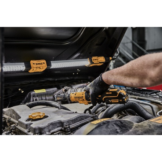A person wearing black gloves uses a DEWALT cordless ratchet tool on a car engine, with a DEWALT work light attached to the open car hood.