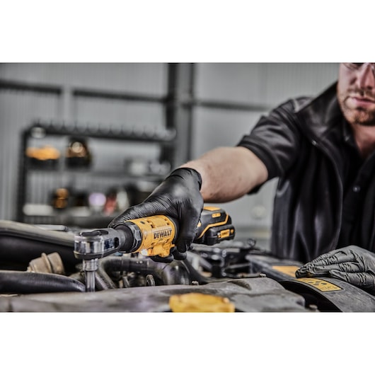 A person wearing black gloves uses a yellow DEWALT cordless ratchet tool (model DCF504L1G) to work on an engine in a workshop.