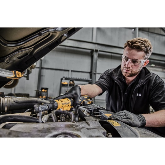 A person wearing black gloves is using a DEWALT DCF504N cordless ratchet tool to work on a car engine in an industrial garage. The face of the person is blurred for privacy.