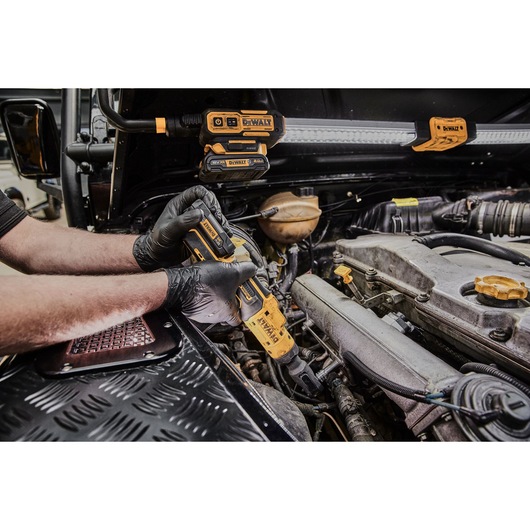 A person wearing black gloves using a DEWALT cordless ratchet tool to work on a car engine in a workshop, with other DEWALT tools visible in the background.