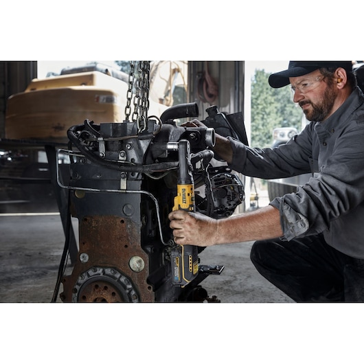 A person in work clothing uses a DEWALT cordless ratchet tool to tighten bolts on a large engine component inside a workshop. The person’s face is blurred for privacy.
