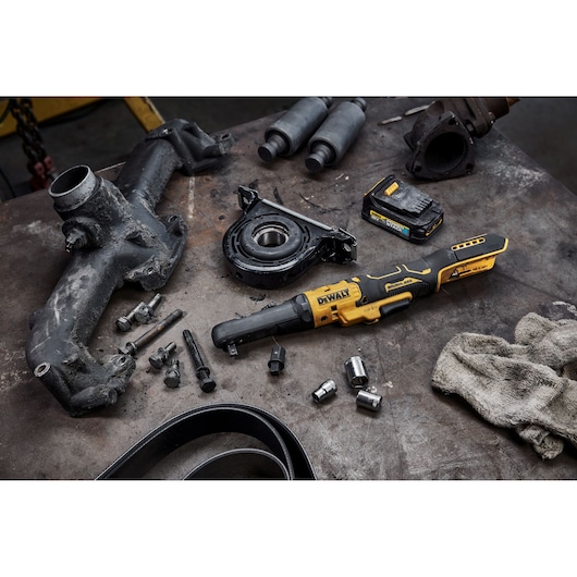 A DEWALT cordless ratchet (model DCF510E2G) is placed on a workbench surrounded by various automotive parts, bolts, sockets, a battery pack, a dirty cloth, and other mechanic tools. The tools and parts appear used, indicating an automotive repair setting.