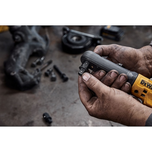 Close-up of dirty hands holding a DEWALT cordless ratchet tool, with various bolts and automotive parts blurry in the background on a workbench.